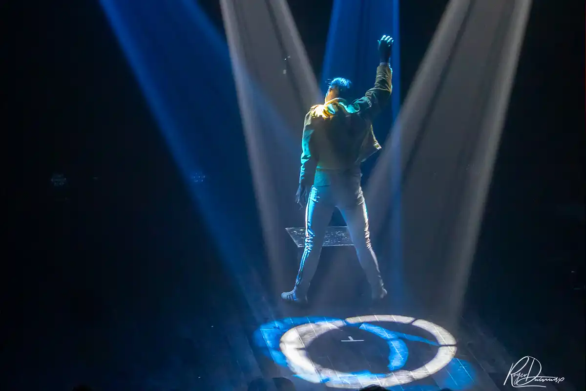 Je Vis avec Freddie Mercury – Ambiance lumineuse Thierry Margot photographié de dos, le bras levé, sous des faisceaux lumineux bleus et blancs sur la scène de L’Esty.
