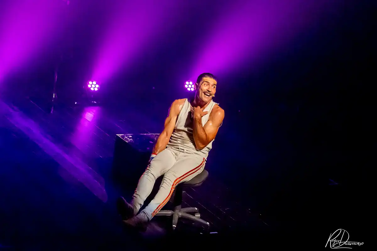 Je Vis avec Freddie Mercury – Scène d’humour Thierry Margot assis sur scène sous des éclairages violets lors du spectacle Je Vis avec Freddie Mercury à Chavanod.