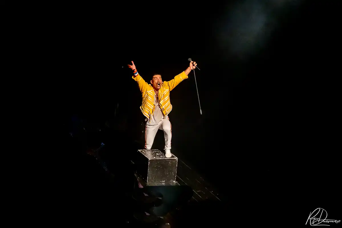 Je Vis avec Freddie Mercury – Thierry Margot sur scène Thierry Margot en veste jaune, debout sur un podium, les bras levés et un microphone à la main pendant le spectacle à L’Esty.